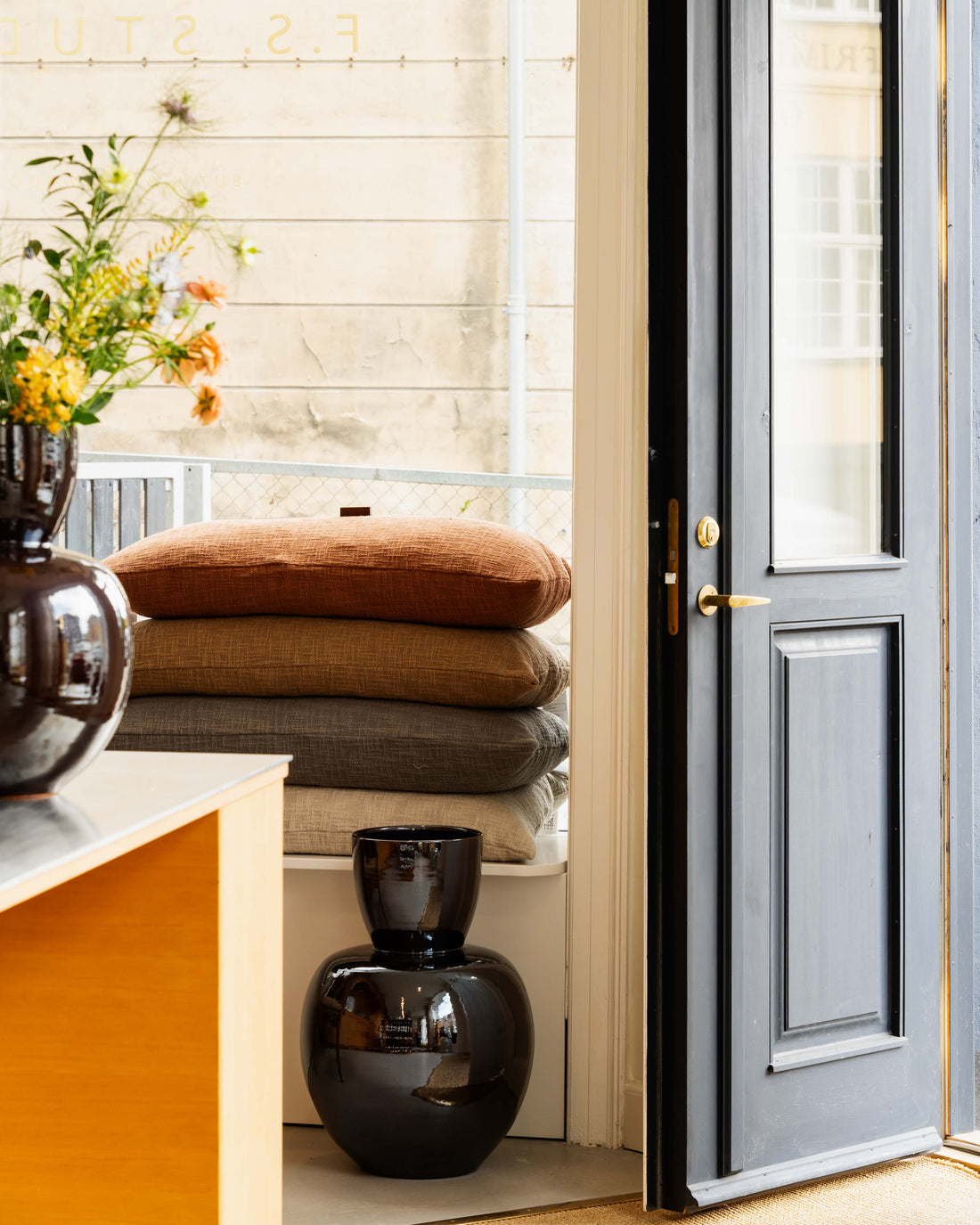 Stack of textured cushions in neutral earth tones by black ceramic vase near open door in TAMEKO fabric store