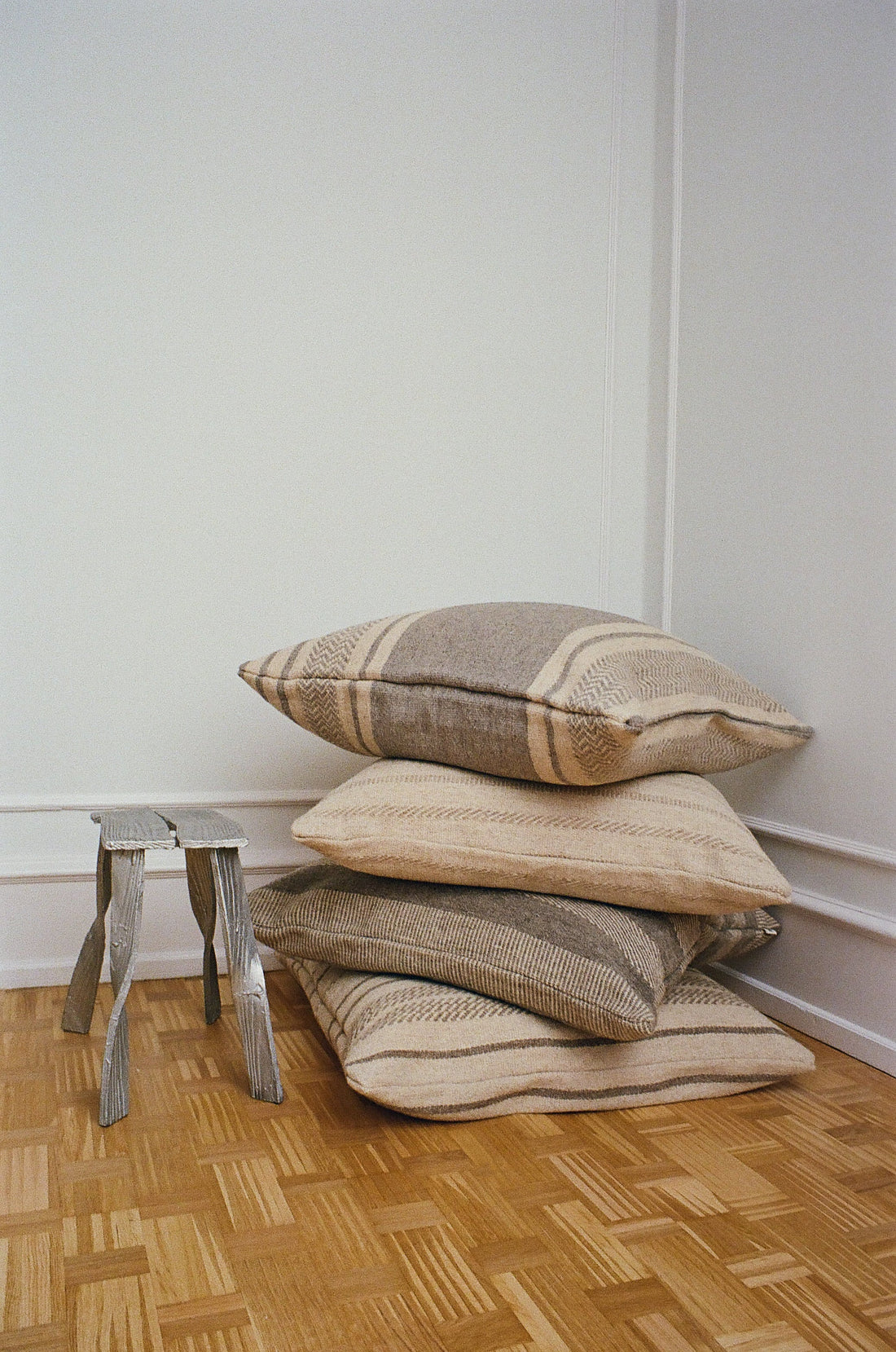 Stack of neutral kelim cushions with subtle patterns on parquet floor, beside small wooden stool, TAMEKO home textiles
