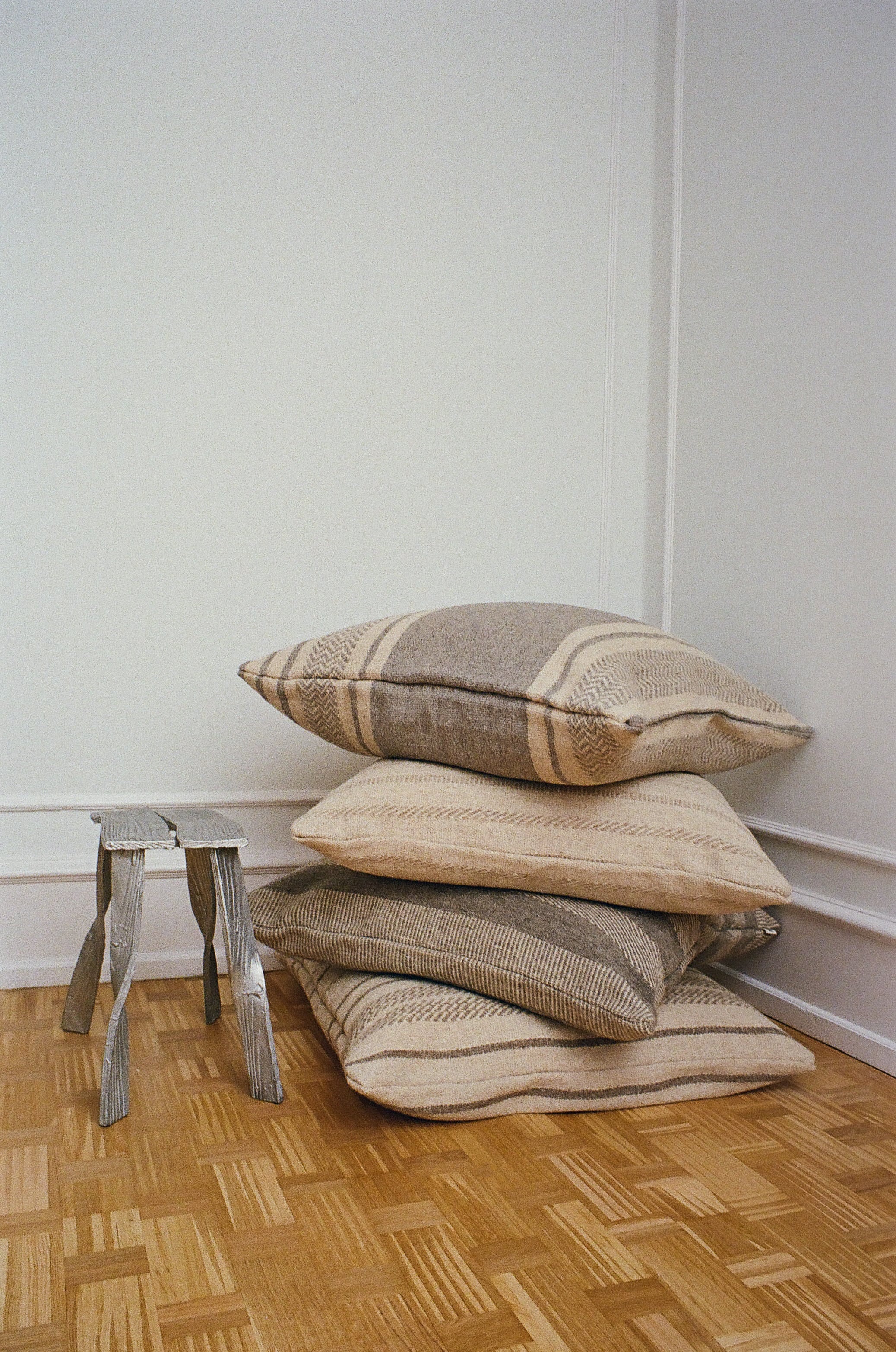 Stack of neutral kelim cushions with subtle patterns on parquet floor, beside small wooden stool, TAMEKO home textiles