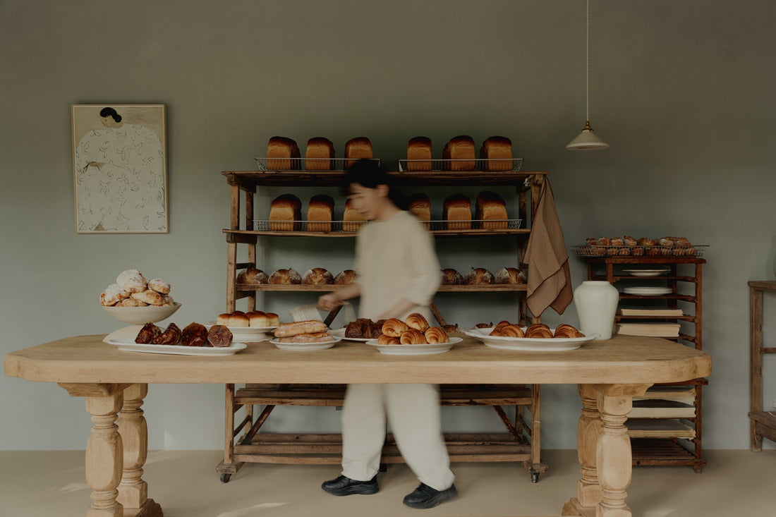 Artisan bakery breads and pastries on wooden table, cozy rustic interior, TAMEKO style.