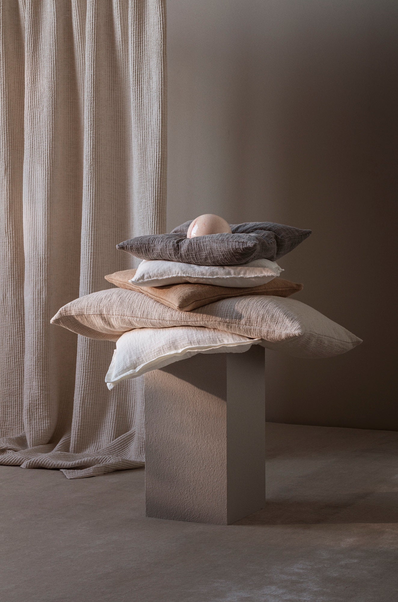 Stack of neutral-toned TAMEKO cushions on pedestal with fabric curtain backdrop