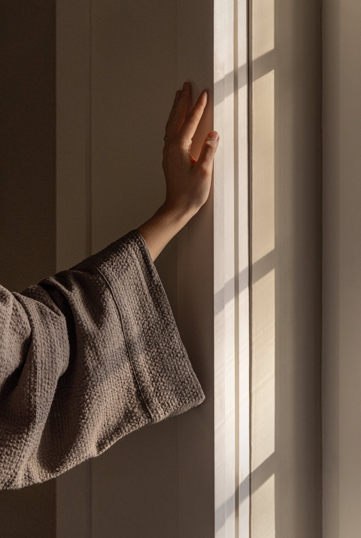 Hand in textured dark kimono sleeve touches sunlit wall, showcasing TAMEKO natural fabric.