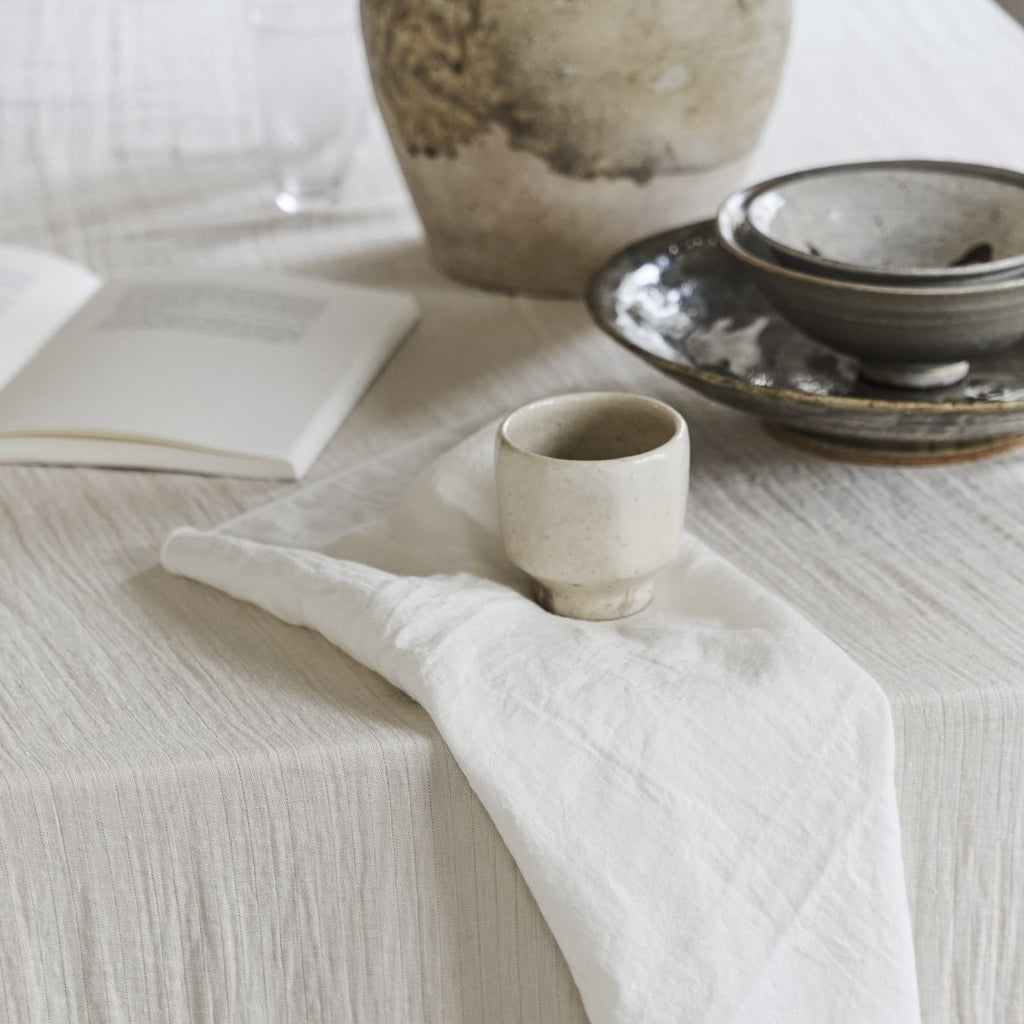Natural textured tablecloth with white Merrow napkin, ceramic cup, and rustic tableware