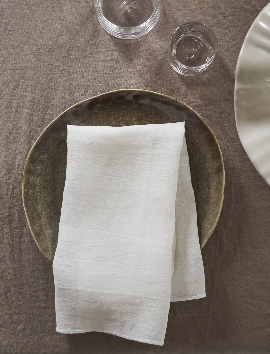 White linen napkin on rustic plate, set on neutral tablecloth, glassware nearby