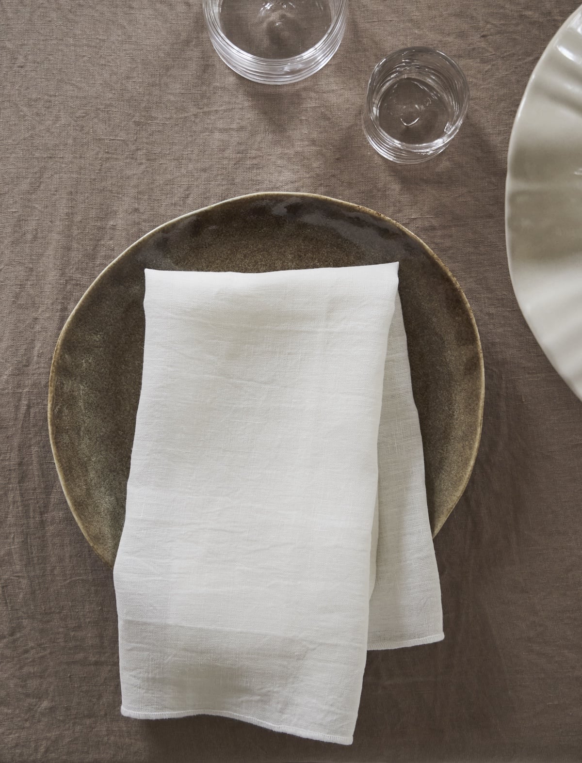 White linen napkin on rustic plate, set on neutral tablecloth, glassware nearby