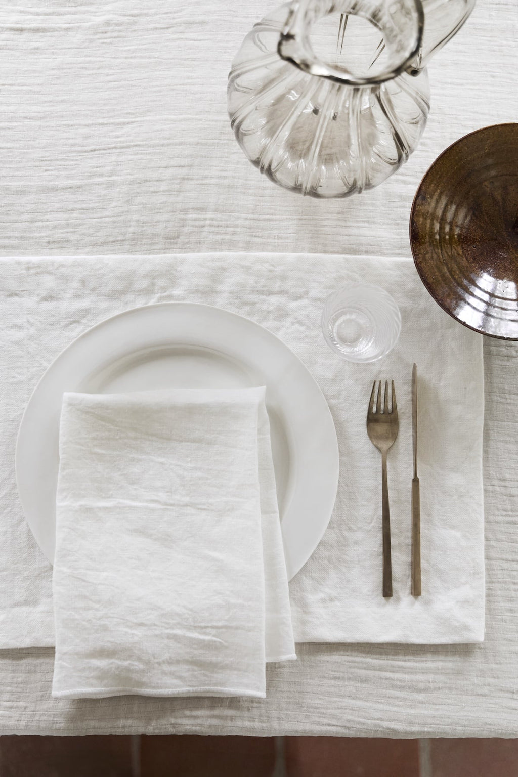 Minimalist table setting with white linen napkin, plate, glass pitcher, and brown bowl on a TAMEKO fabric tablecloth.